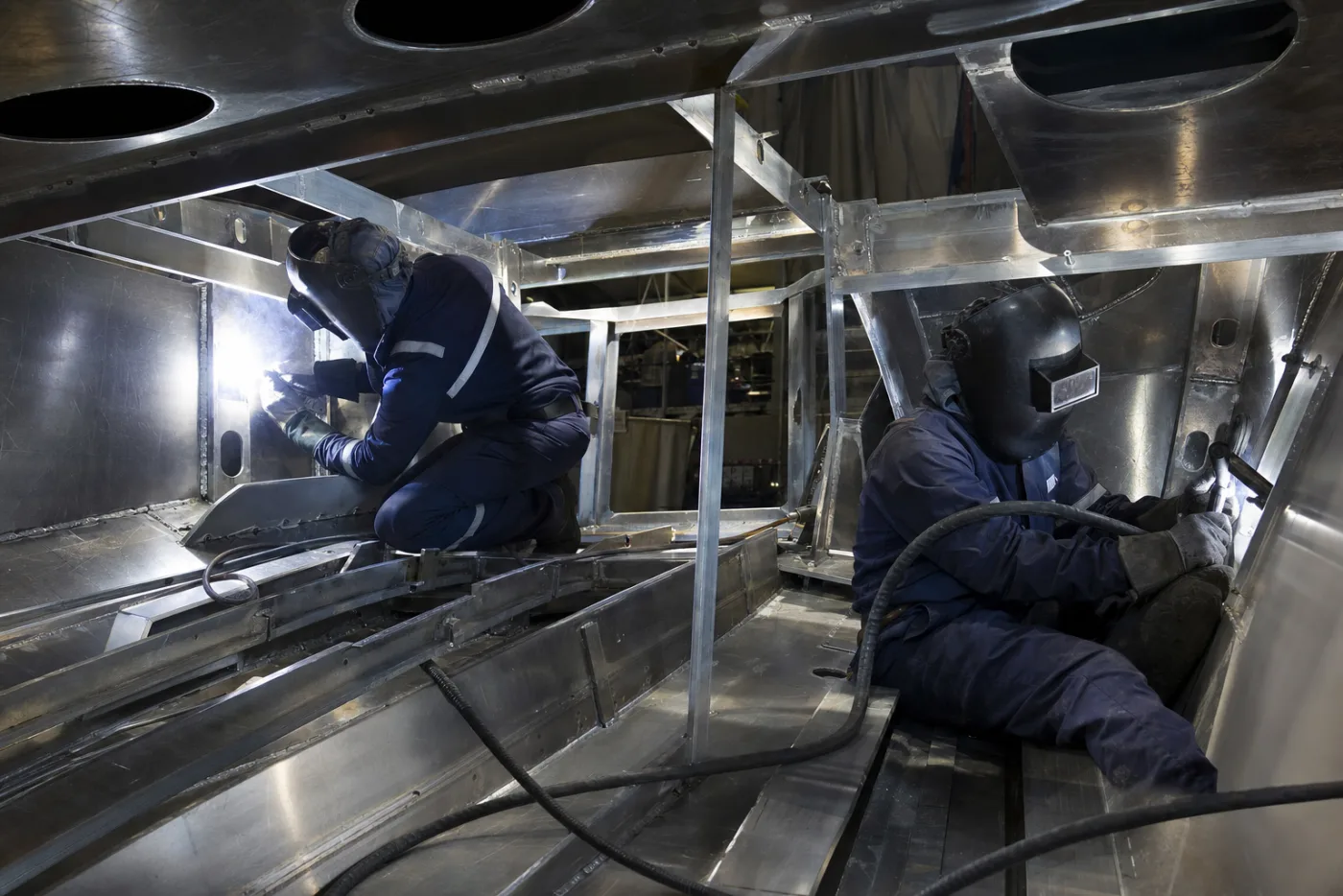 A bare aluminium hull under construction in a covered hall